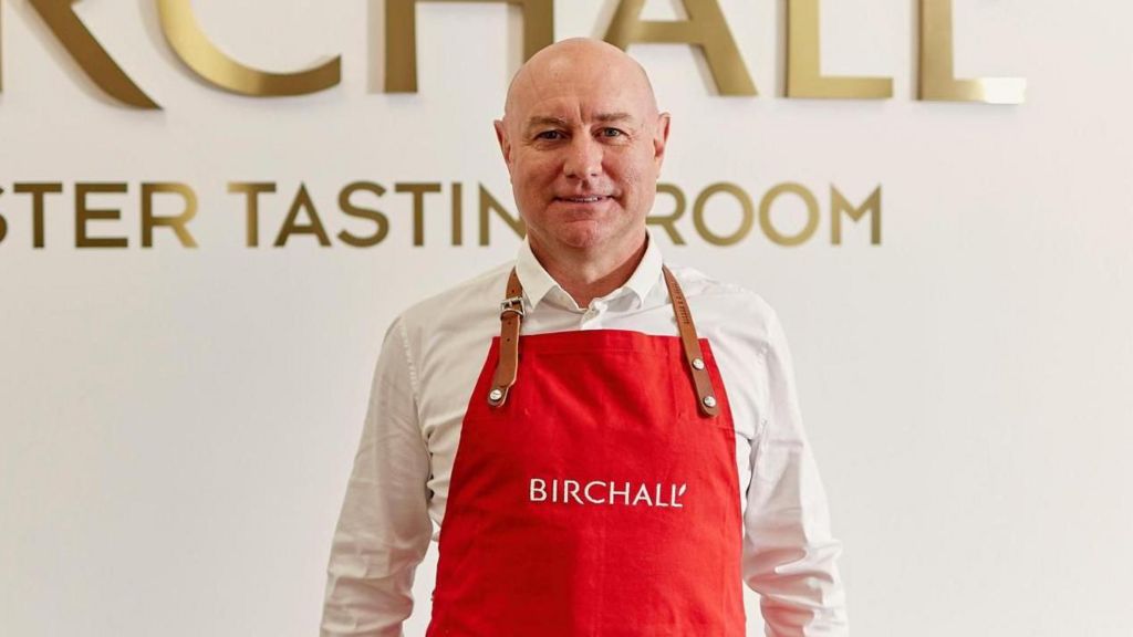 Daniel Graham, wearing a Birchall-branded red apron and white shirt, standing in front of a white wall with the words BIRCHALL MASTER TASTING ROOM written on it in gold letters