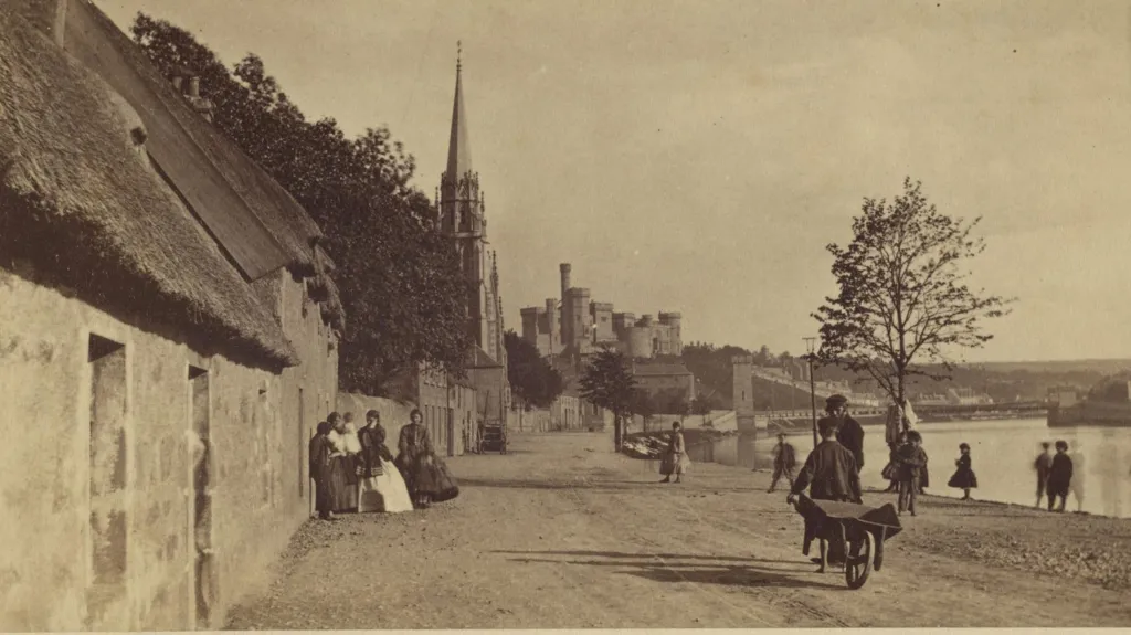 A sepia photograph of Inverness riverside with a row of cottages, a church and in the distance the hilltop castle. A boy in a cap and pulling a barrow is talking to a man wearing a cap. A group of women and children stand by the cottages and other people are standing near the river.