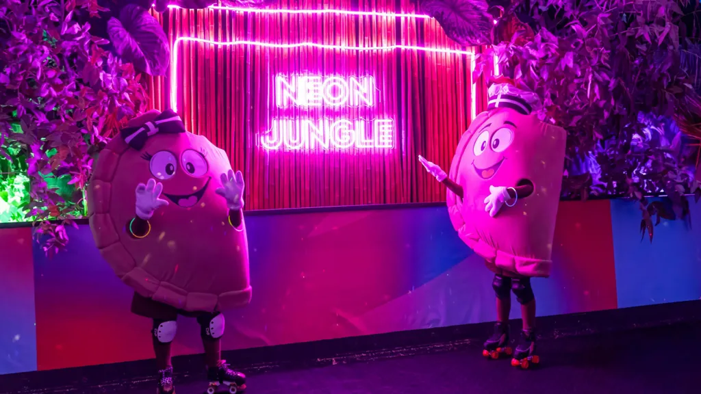 Two individuals dressed in a pasty costume with roller skates on, stood in front of a neon sign which says 'Neon Jungle' on it.  There are two faux leafy bushes, one in the top right and one in the top left. One of the individuals is wearing a bow on their costume which is a Cornish Flag design, and the other is wearing a chef's hat with a Cornish Flag design on too.
