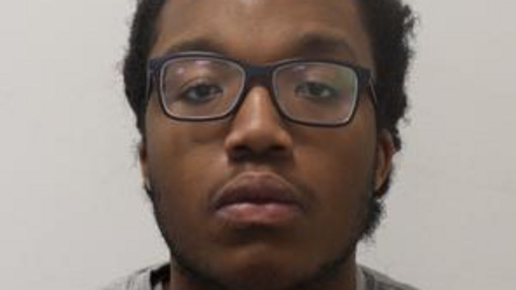 A police mugshot of a young man wearing dark-framed glasses