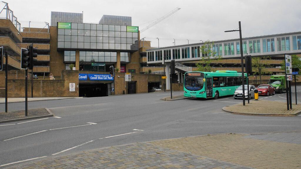 Cavell Blue Car park, view from other side of the road, also showing a teal-coloured bus and three cars waiting in traffic and the footbridge that leads from the car park to the shops