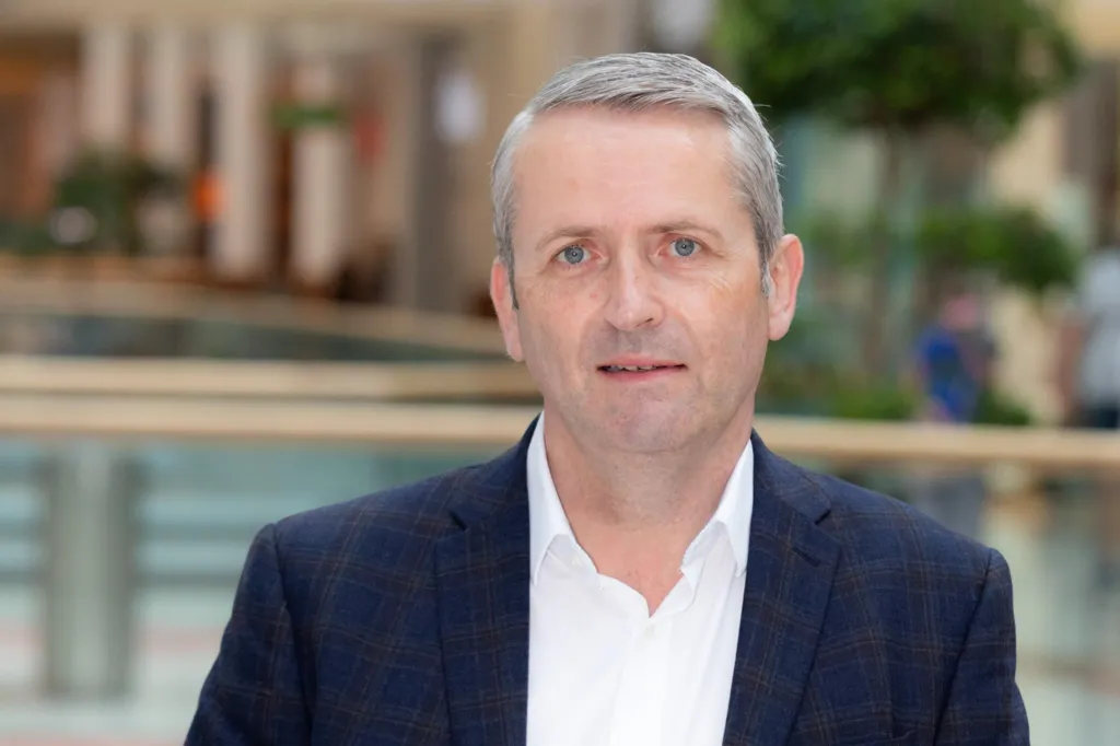 Jonathan Poole has short grey hair and is wearing a white open-neck shirt and a dark suit jacket