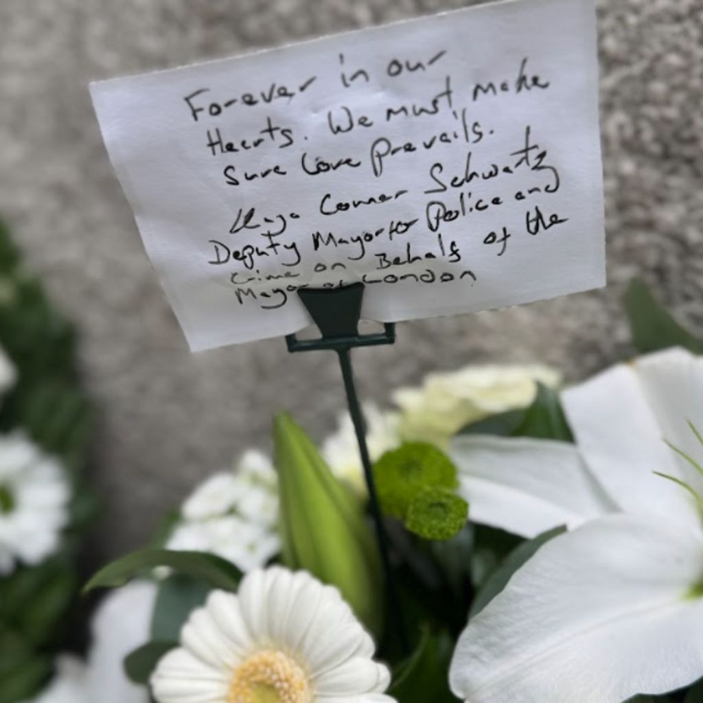 A handwritten note reading “Forever in our hearts. We must make sure love prevails,” placed among white flowers in a memorial arrangement.