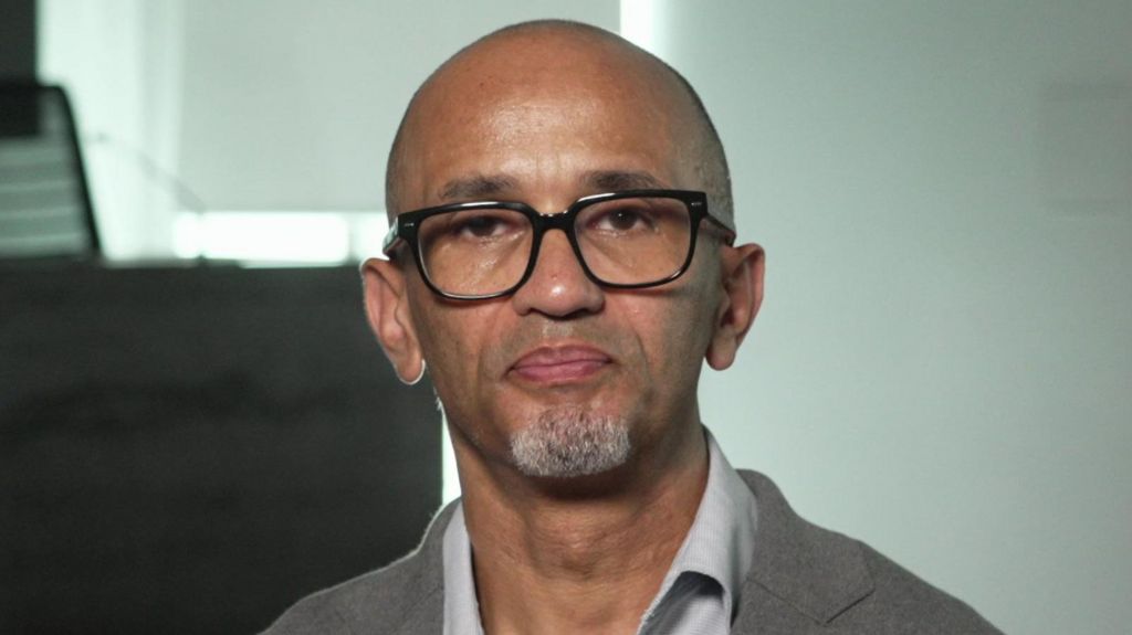 Desmond, now in his fifties, looks directly into the camera. He is wearing glasses and a light grey suit jacket and shirt.