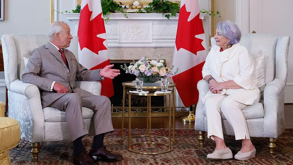 An image showing Mary Simon sitting across King Charles III at a formal meeting. Behind them are two Canadian flags. Simon is wearing a white suit, and she has grey and lilac hair. King Charles III is wearing a grey suit with a red tie. They are sitting on white arm chairs, and behind them is a white fireplace. 