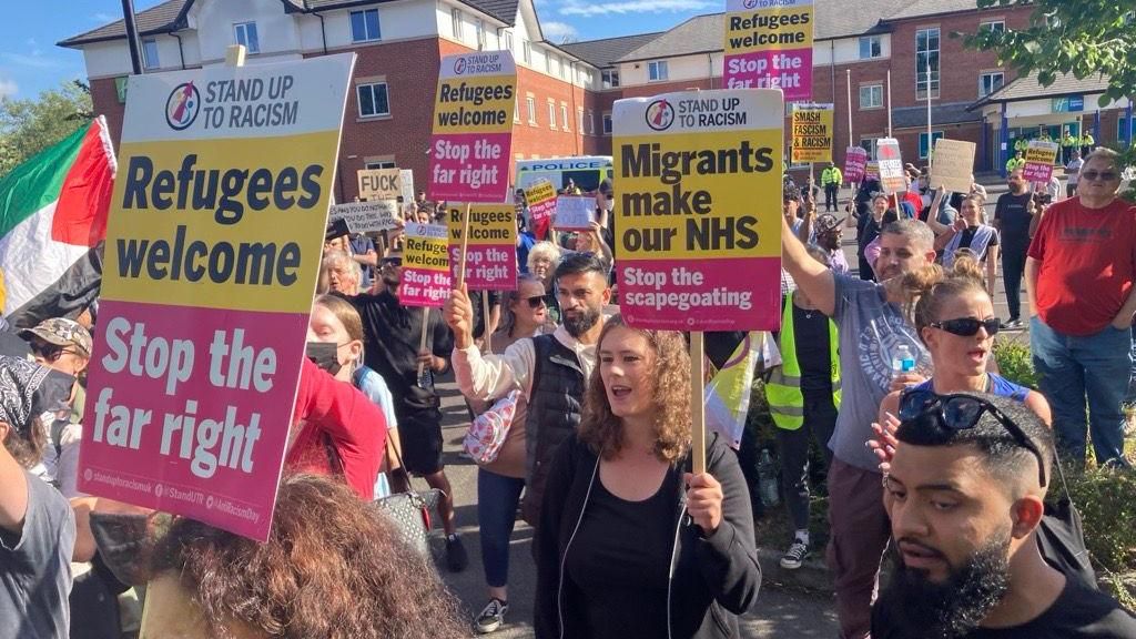 A crowd of chanting men and women carry placards with slogans such as "Refugees welcome" and "Migrants make our NHS" at a counter-protest in Crawley