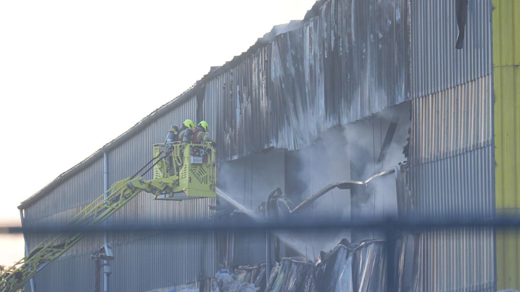 Two firefighters in high-visibility protective gear stand in the basket of a turntable ladder, directing a water jet into a large hole in the charred, corrugated metal wall of an industrial building. Smoke billows from the damaged structure.
