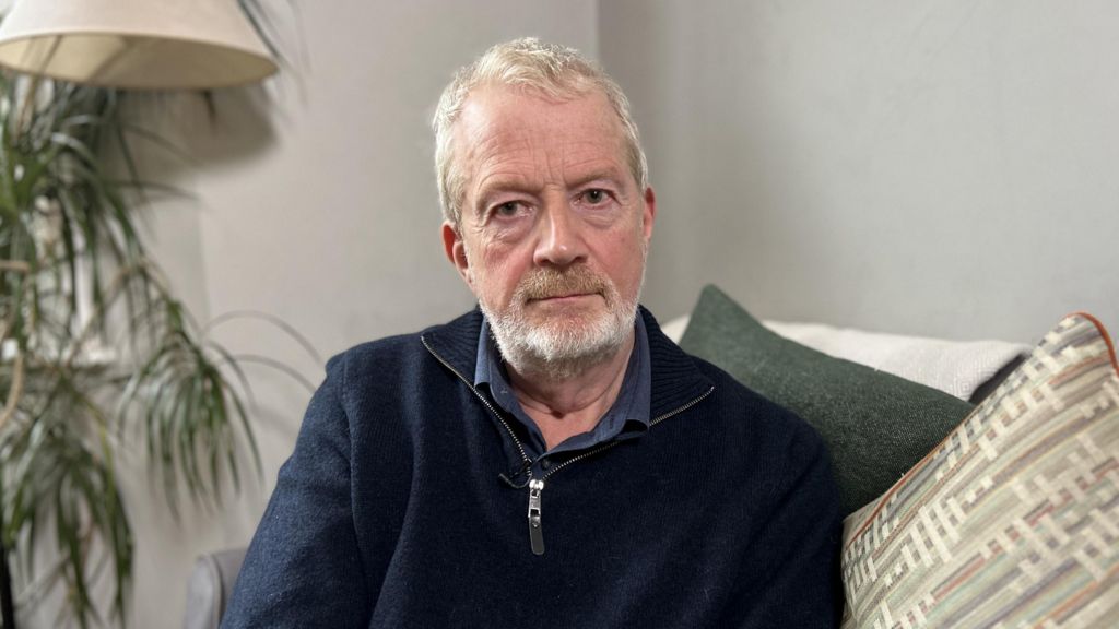 A man with short grey hair looks at the camera. He is seated, with cushions behind him and to his side. He is wearing a dark blue fleece with a zip, which is open at the neck