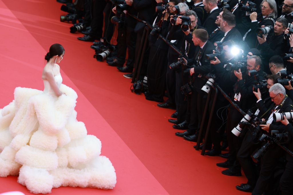 An actress in an elaborate voluminous white dress on the Cannes red carpet facing a bank of photographers in black tie.