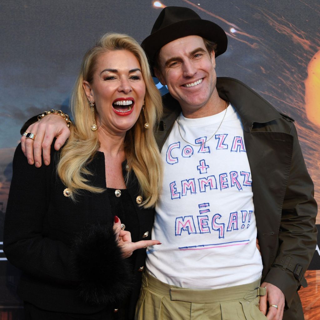 Claire Sweeney and James Cartwright smile and stand together in front of Corriedale backdrop at the premiere. He has his arm around her shoulder and he's wearing a white T-shirt with a DIY slogan written in pink and white. She's laughing and pointing at it.