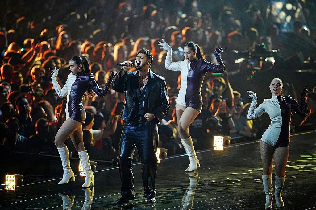 Noam Bettan singing in a black outfit with three women dancing around him in black and white short outfits and white boots