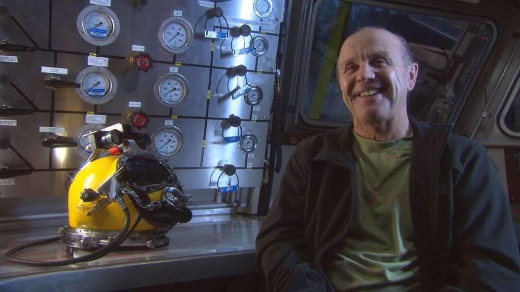 Doug Allan smiling at the camera. He is sitting down and is wearing a dark, open jumper over a green t-shirt. A yellow divers helmet is on the table next to him.