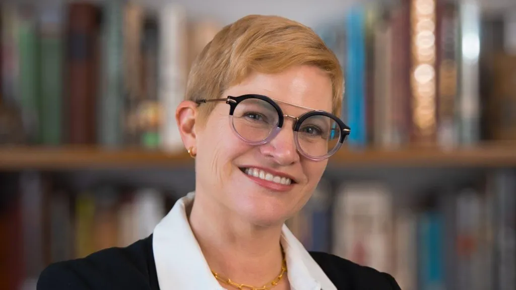 A head and shoulders portrait of a woman with short hair and glasses, smiling. She is wearing a black jacket and white shirt and stands in front of bookshelves, which are in soft focus.