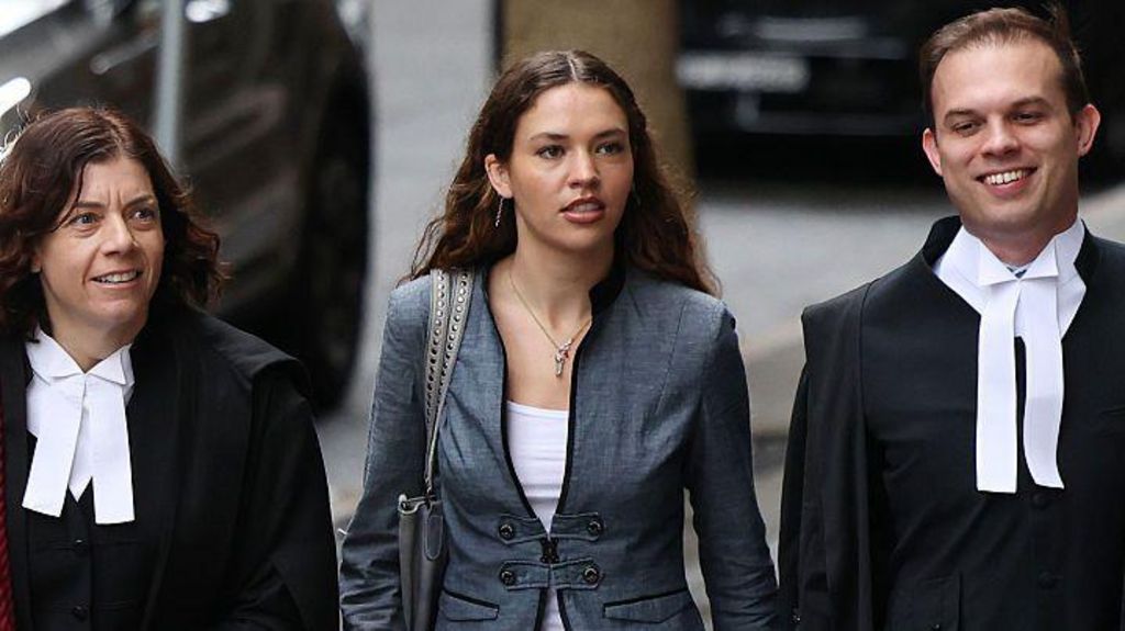A young woman with long brown hair, wearing a white top and grey jacket, flanked by two barristers in black and white legal gowns, walking down a street