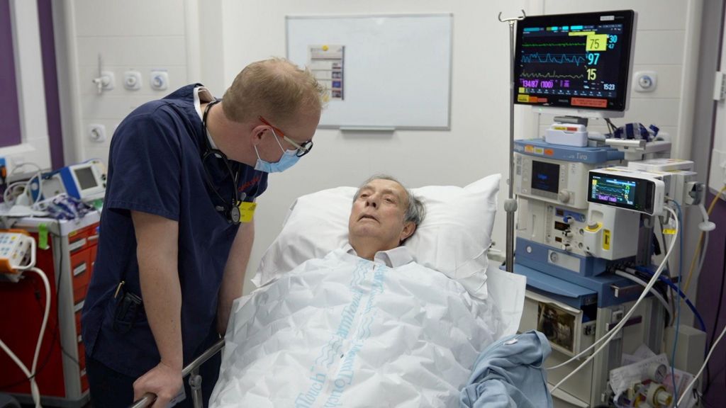 A man lays in a hospital bed with a nurse speaking to him.