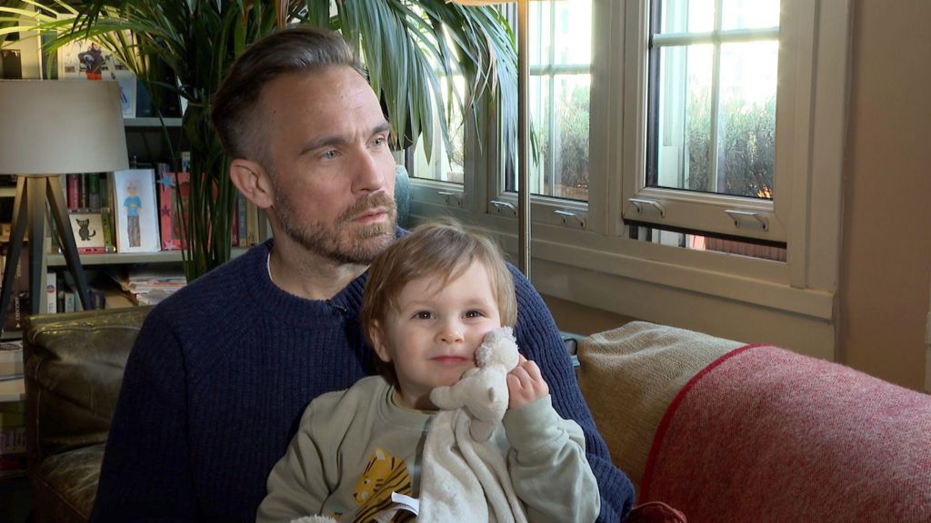 The Dad holds his son who clutches a teddy to his face. They sit on a sofa with a plant in the background.