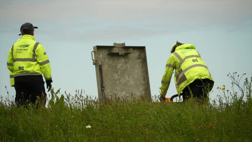 Two men wearing South West Water high-viz uniform are in a field. One is looking into a large metal open drain cover. The other is standing nearby.