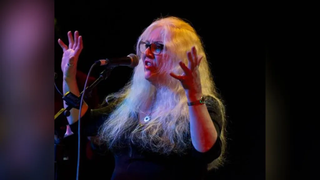 Alison is onstage. She's singing in front of a mic with her hands up. She wears glasses and has long hair. She's wearing a silver necklace and a watch.