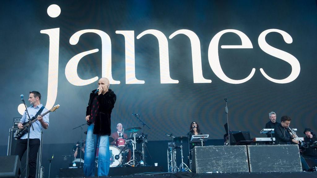 A large stage backdrop reading "james" in white letters. on stage are the band james. you can see one member in a blue shirt playing the bass guitar, next to a bald man singing into a microphone. in the background are two drummers, a man on a keyboard, a man playing a trumpet and a photographer.