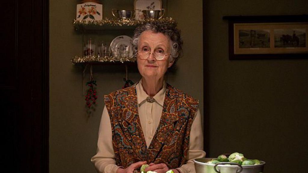 Miss Higgins, played by Georgie Glen, peeling sprouts in the kitchen in a white blouse and apron