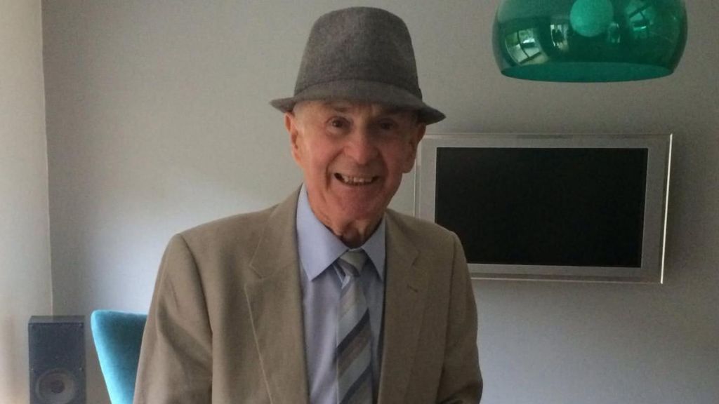 An elderly man smiles for a photo. He is wearing a grey hat and a cream jacket and light blue shirt.