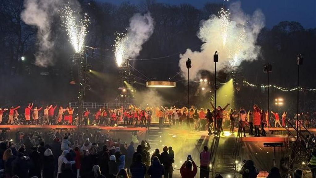 A crowd of people watching on as dozens of people perform on a stage. There is various stage equipment such as standing speakers and plumes of smoke being let off.