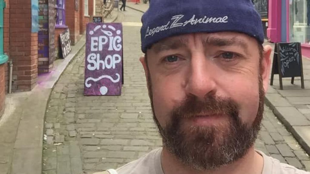 Ben Gunnery has a goatee beard and is wearing a purple beanie hat.  He is taking a selfie photo in a narrow street full of shops.