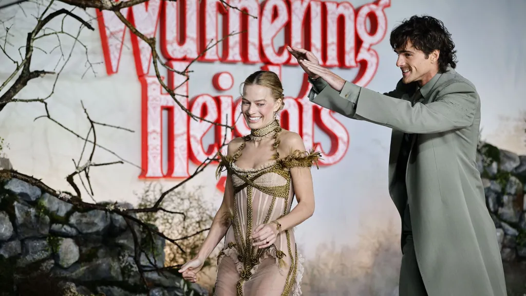 Jacob Elordi puts his hands above Margot Robbie's head in an attempt to shield her from the rain as they both smile.