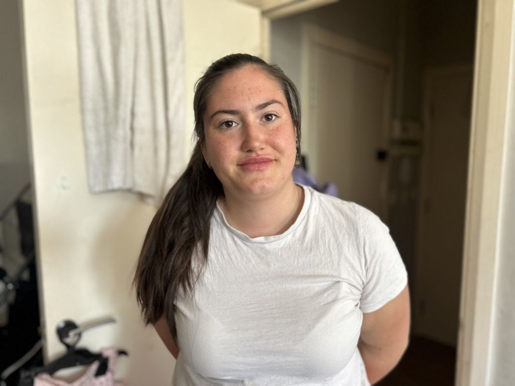 Woman with long brown hair tied in a ponytail to one side, and wears a white t-shirt. She is standing in a light-coloured room.