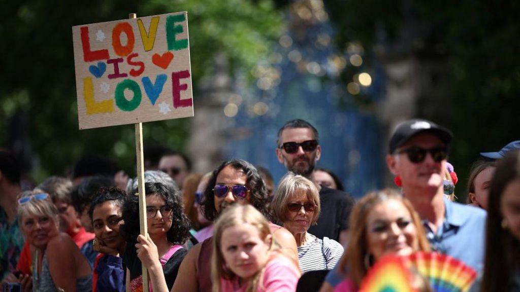 A member of the public watching the parade holds a placard with rainbow letters saying "Love is love". They are surrounded by other people all looking in the same direction