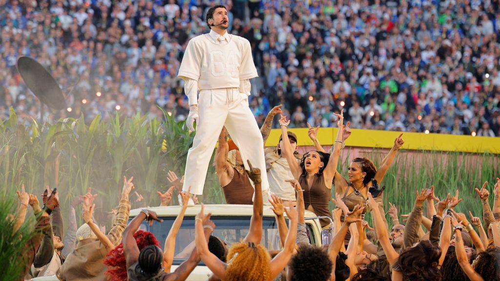 Bad Bunny on stage at the Superbowl. He is standing on a car and surrounded by female dancers waving their arms. 