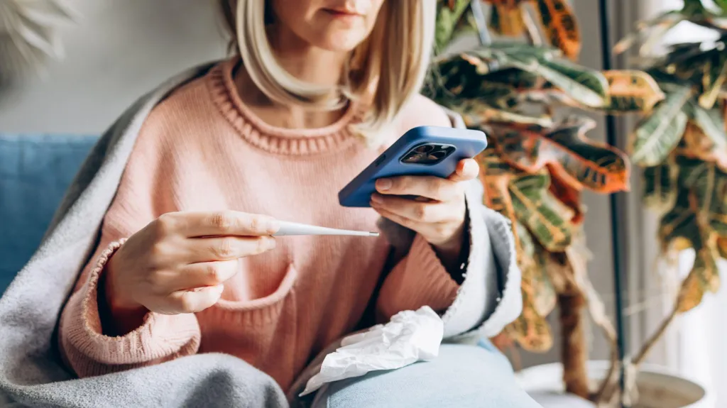 Sick woman at home wrapped in blanket measuring temperature and talking on smartphone for medical consultation.