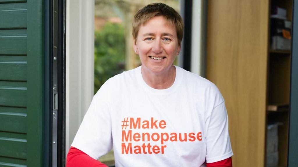 Diane has short, brown hair and is wearing a white tshirt with the words "Make Menopause Matter".