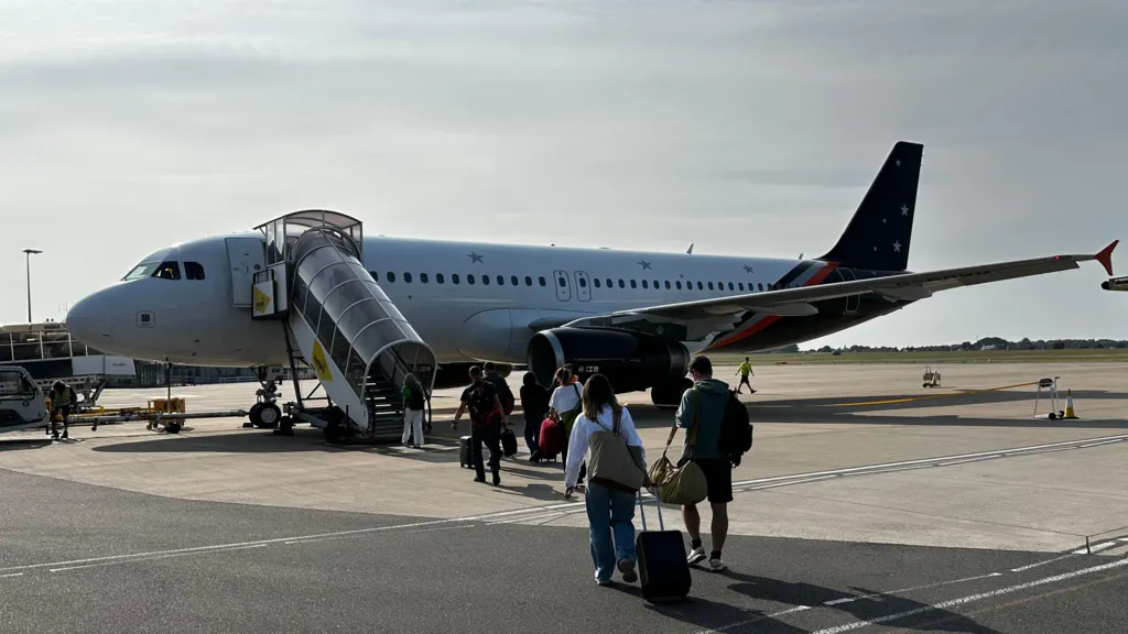 A Titan aircraft, with passengers going up a glazed stairway. It's a stubby jet aircraft with a white body and a blue wing and tail. 