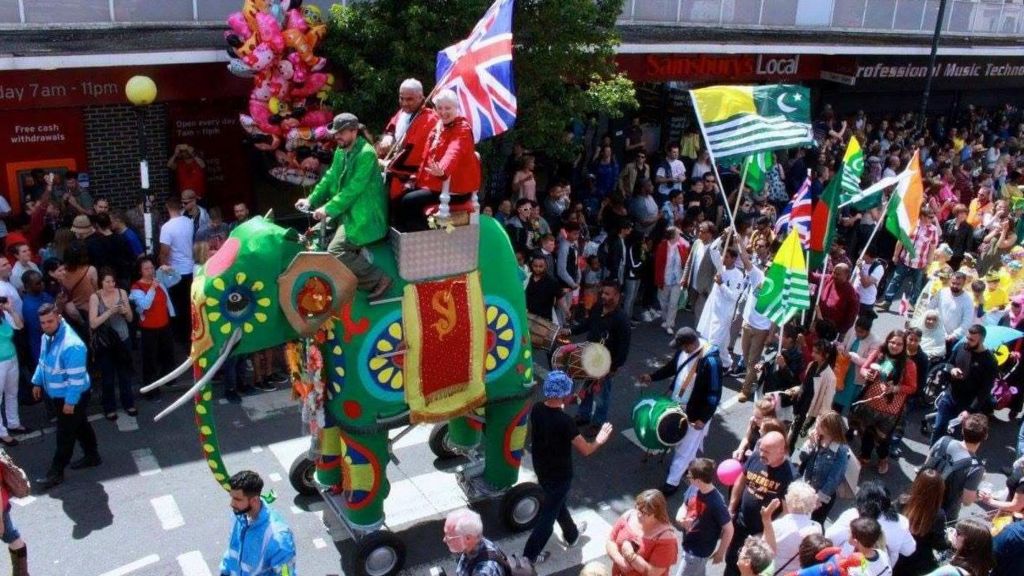 People at a street carnival, dressed in bright colours, watching a green elephant floats parade through the streets