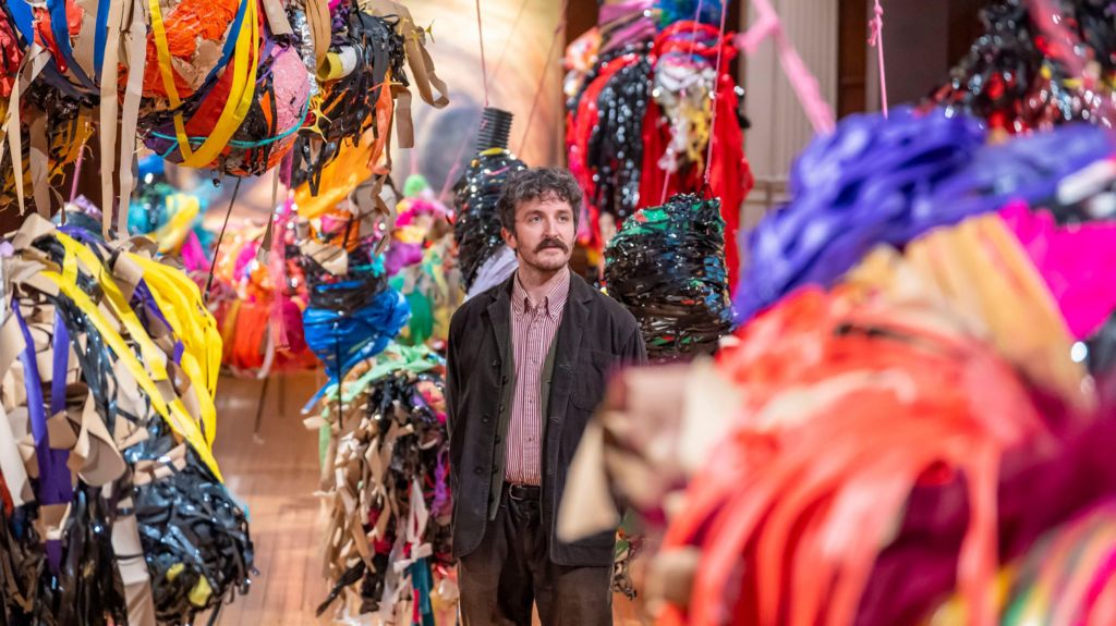 A man standing in the centre of a large number of suspended colourful sculptures made of various strips of multi-coloured tape and material in Nnena Kalu's installation