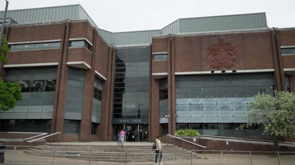 A large building with brown brick and glass panels. Steps lead up to the entrance of the building with a handful of people outside. 