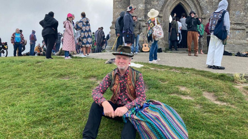Mike Daniels sitting on the grass in front of Glastonbury Tor wearing a patterned shirt, waistcoat and hat 
