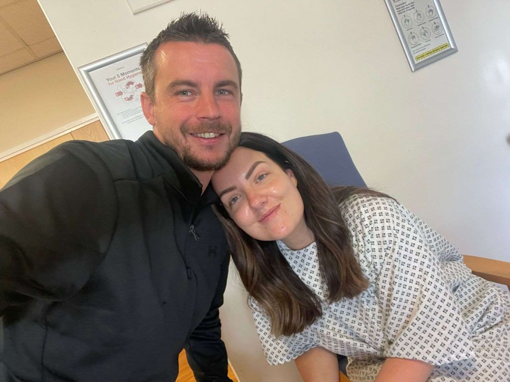 Photo of lady in hospital gown in a clinical area of a hopsital with her partner, a man dressed all in black. Both are smiling at the camera.