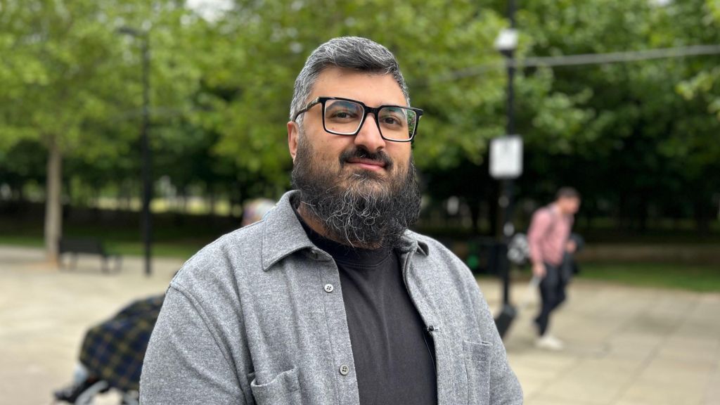 A man wearing glasses and a grey shirt over a black top standing outdoors near a park, with trees, a paved walkway, and a passer-by in the background