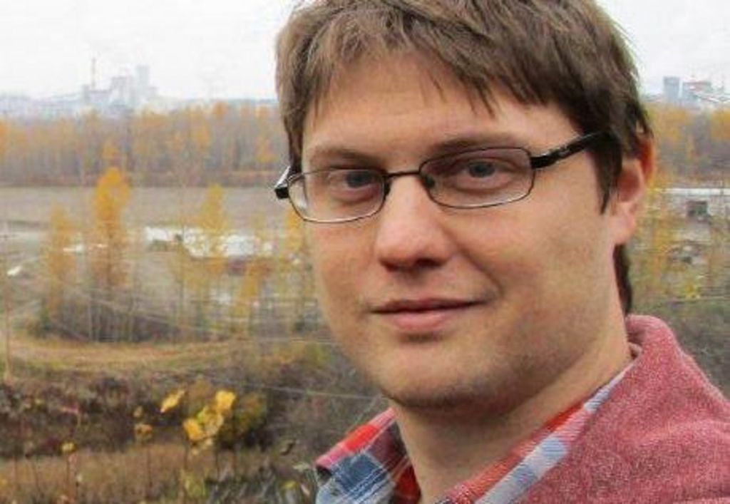 Peter Todd looks at the camera while wearing glasses and a red shirt and standing outside with autumnal trees and a power station in the background