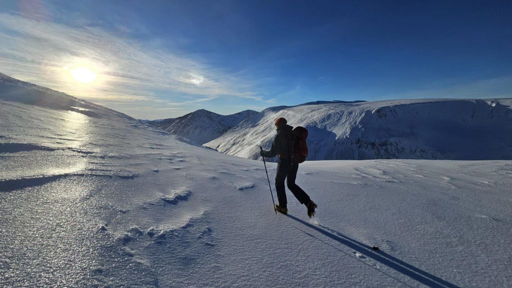 A single walker crosses the snow and ice on the way to the top of the mountain, Ben Macdui in the Scottish Highlands. They are walking towards the sunshine and the whole countryside is covered in white snow.