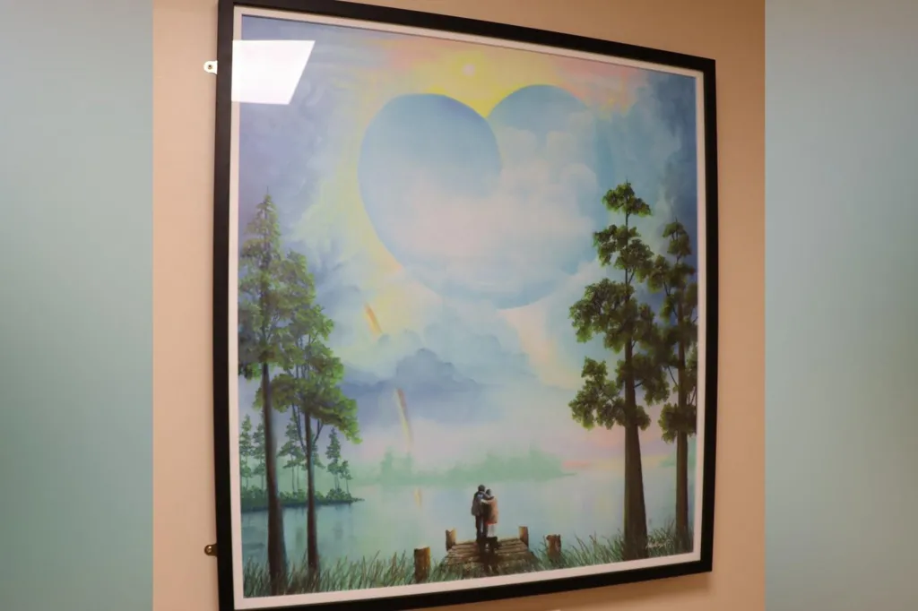 The pastel artwork is of two figures arm-in-arm at the end of a wooden pier. The sky above the figures is blue, and is dotted with clouds including a heart-shaped one. A rainbow breaks through the cloud and is reflected in the water below. The scene is bordered by tall trees.