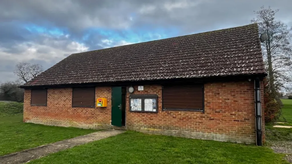 A one storey building pictured from the outside surrounded by grass. It is made of red bricks and has a green door. The clouds above are grey.