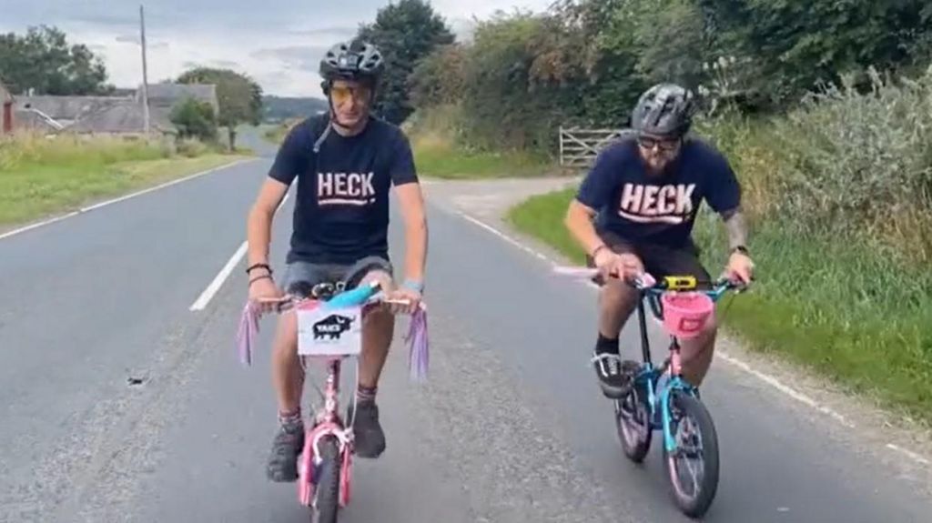 Adam Alderson rides a pink children's bike on a North Yorkshire road while wearing a cycle helmet and a black T-shirt with the branding 'Heck' across the front. His friend can be seen riding next to him in similar fashion