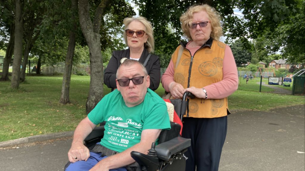 A man in a wheelchair in a green top and glasses in front of two women with blonde hair.