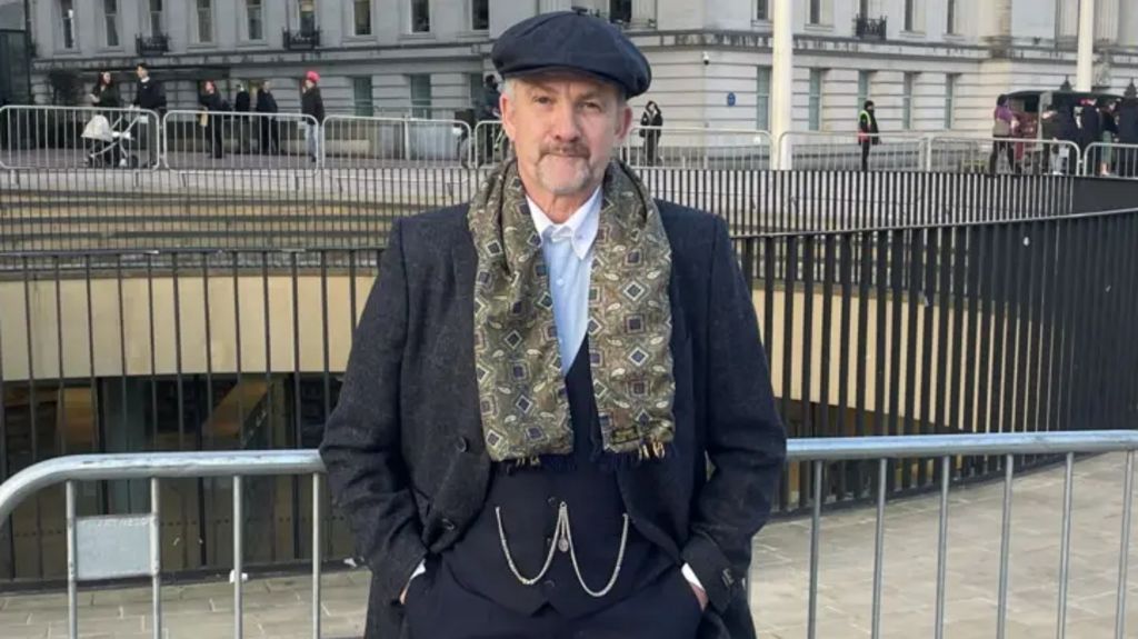 Arthur Cocker, a man dressed in a dark-coloured waistcoast and jacket with a white shirt and a dark-coloured flat cap with a patterned scarf. He is standing by railings in a city centre.