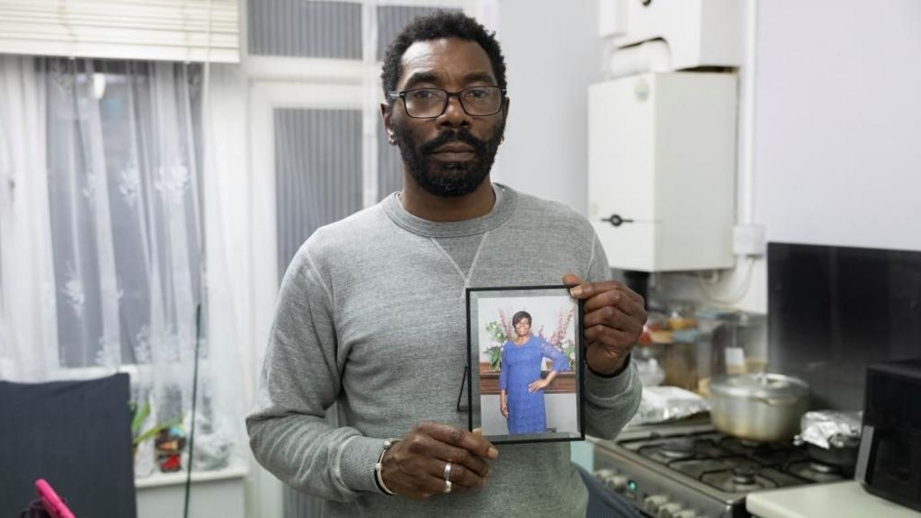 A man in a grey jumper hold photo of his mum in a blue dress. There is kitchen in the background where the hob can be seen.