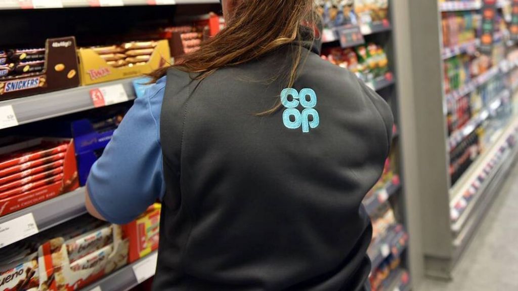 A woman with a long brown ponytail wears a black gillet with a blue co-op logo while looking at a shelf full of chocolates in the supermarket.