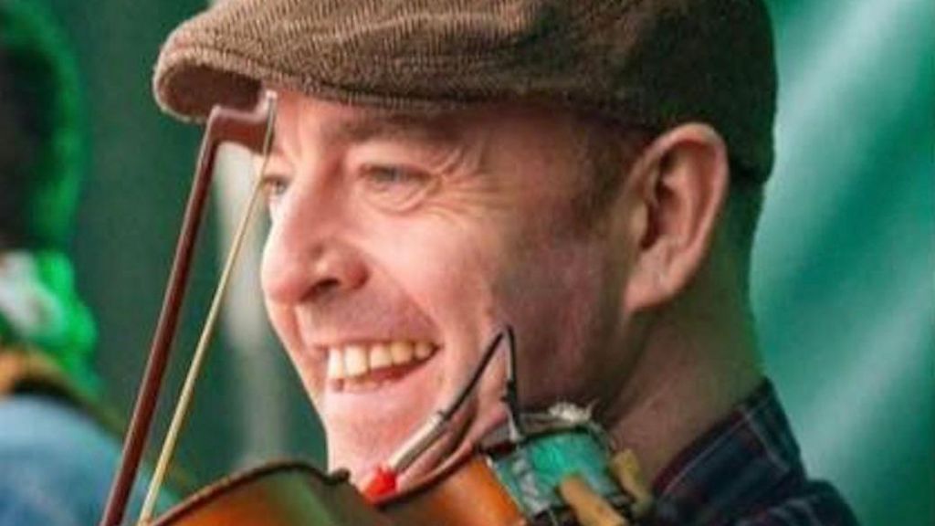 Ben Gunnery is playing the violin and smiling.  He is wearing a peaked tweed cap and a checked shirt